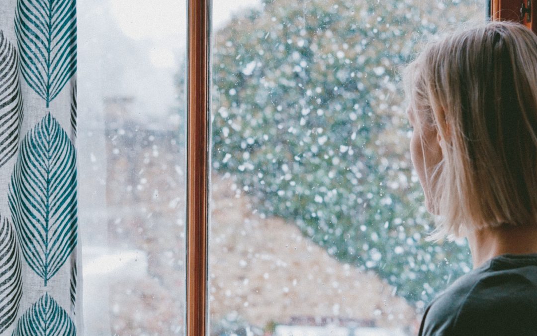 A woman stands inside, looking outside a window where it is snowing. She is holding a mug.
