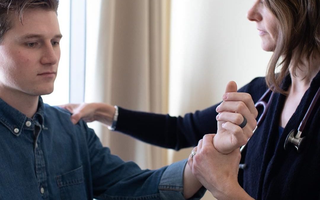 A female integrative primary care practitioner examines a patient's shoulder. She is holding his left wrist, and is examining his left shoulder.