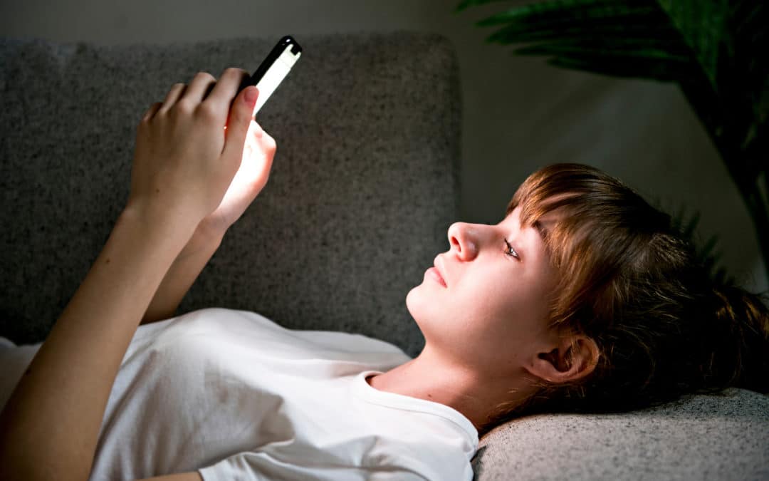 A teen girl is lying on a sofa and holding her phone. She is looking up at the screen, which is shining a white light on her face.