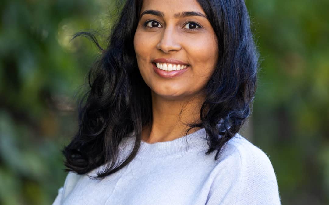 Dr. Aneela Cox smiles, facing the camera, with trees blurred in the background.